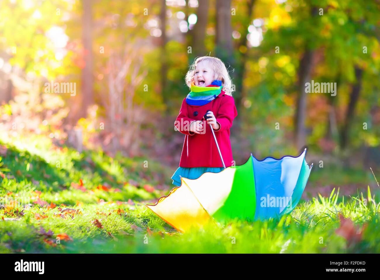 Little girl playing in the rain in autumn park. Child holding umbrella walking in the forest on a sunny fall day. Stock Photo