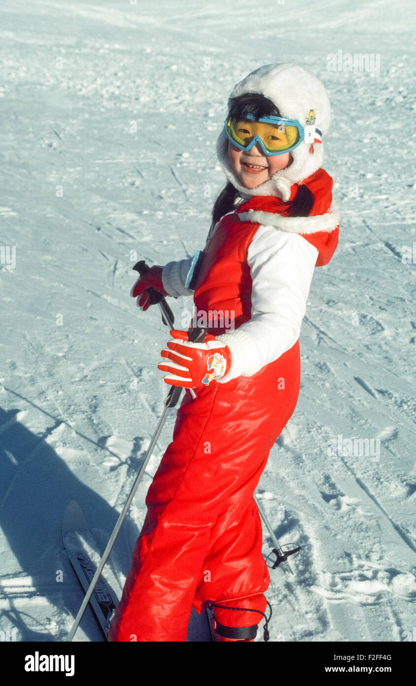 A cute young Asian girl in a bright red ski suit, blue ski goggles and furry white ski hat smiles in excitement as she is ready to descend a bunny slope on Mt. Alyeska in Alaska, USA. Stock Photo