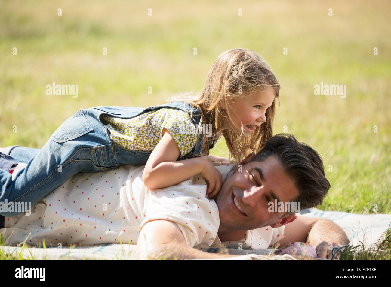 Playful daughter laying on top of father in sunny field Stock Photo