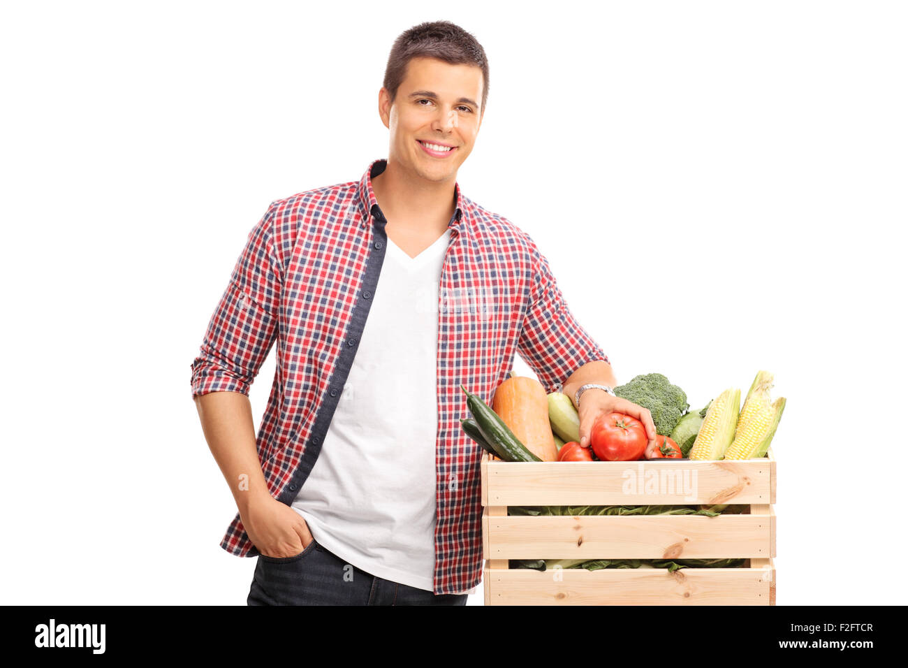 Young man leaning on a crate full of fresh vegetables and looking at the camera isolated on white background Stock Photo