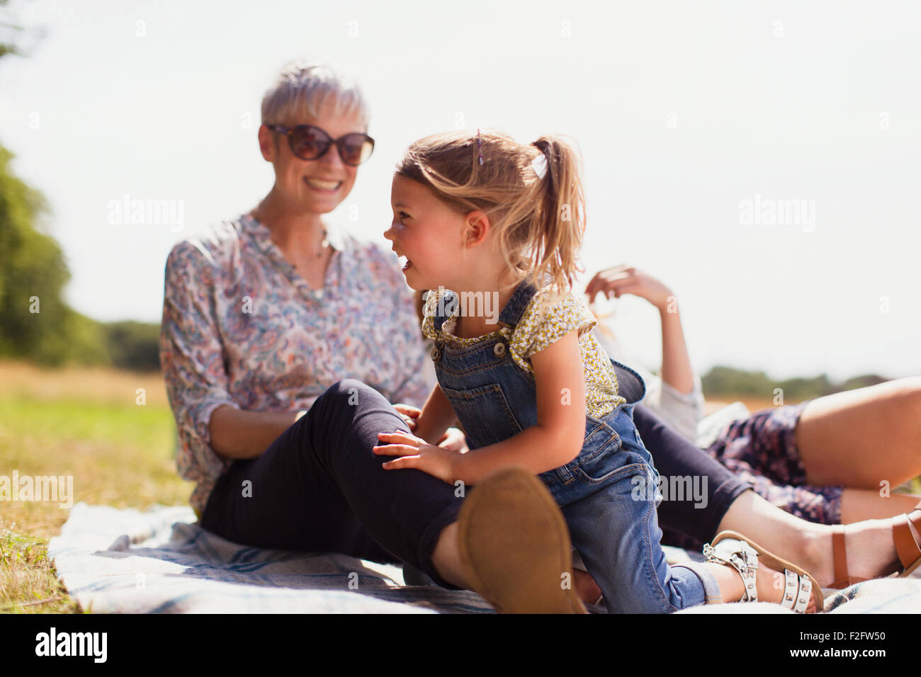 Grandmother and granddaughter laughing on blanket in sunny field Stock Photo
