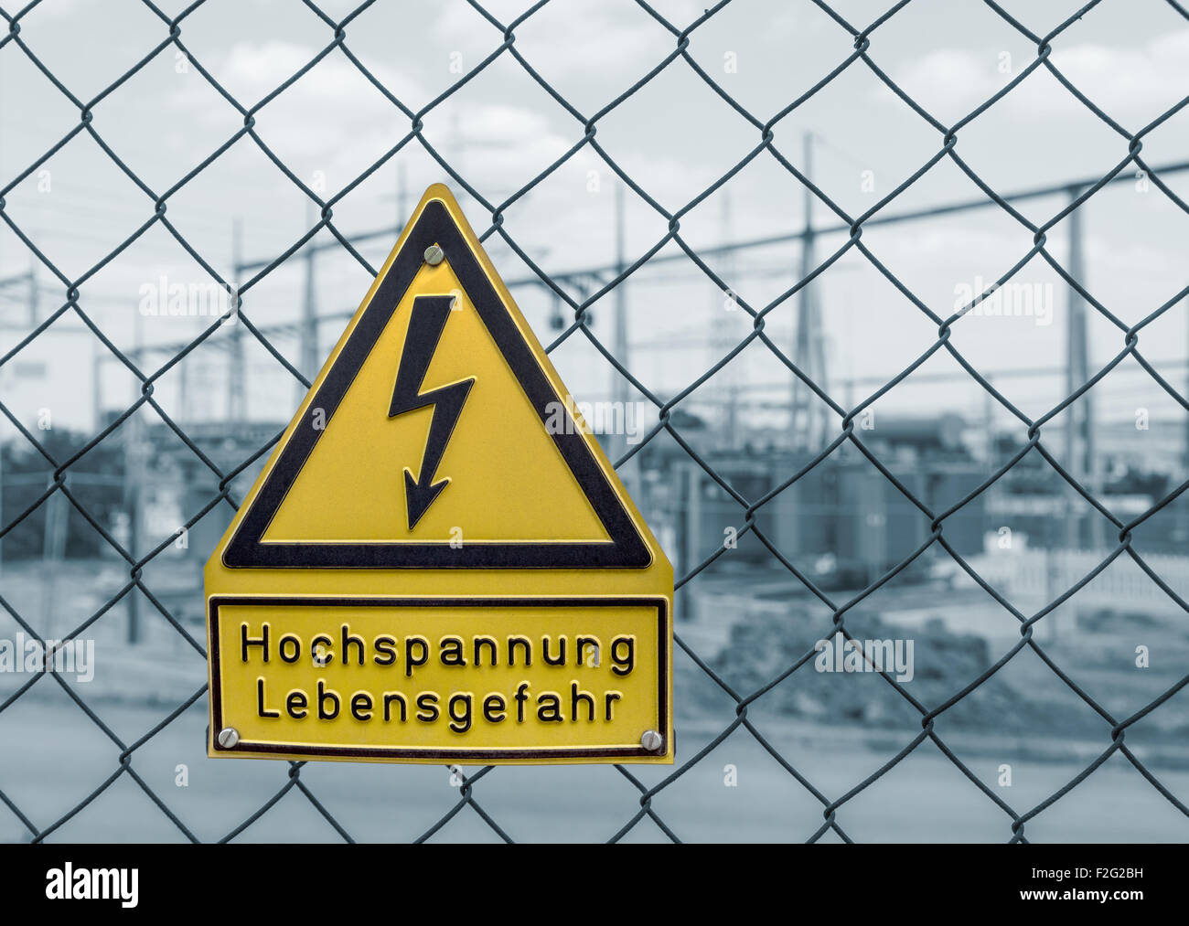 yellow german high voltage warning sign on a mesh wire fence in front of a blue toned electrical substation in Southern Germany Stock Photo