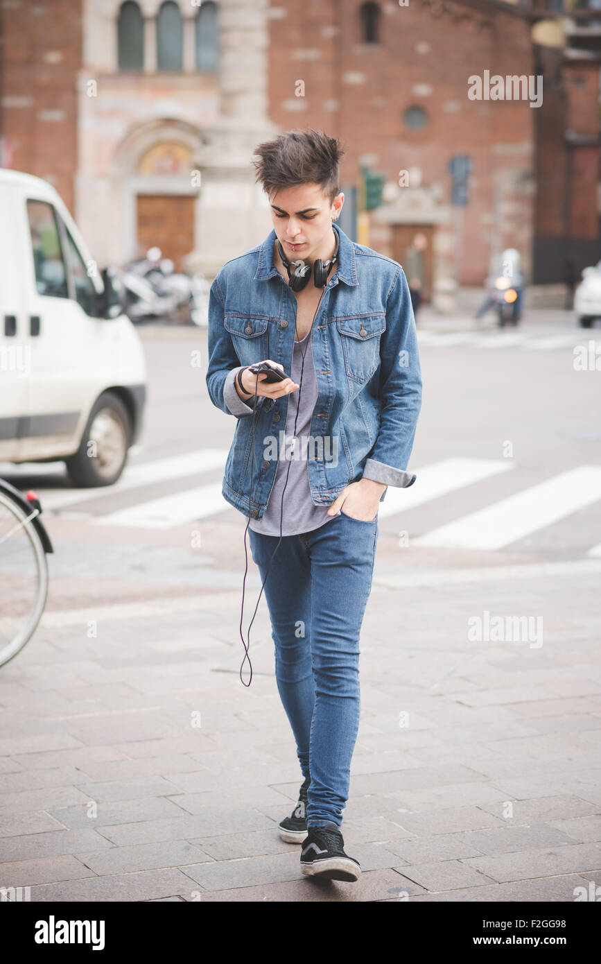 young handsome alternative dark model man walking trough the street listening to music with headphones and smartphone connected Stock Photo