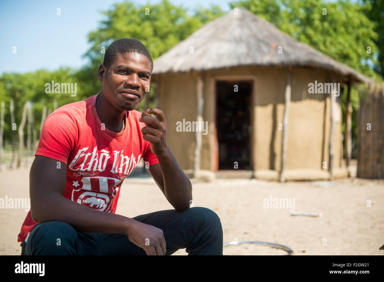 African man sitting in front of thatched roof hut in Botswana, Africa Stock Photo