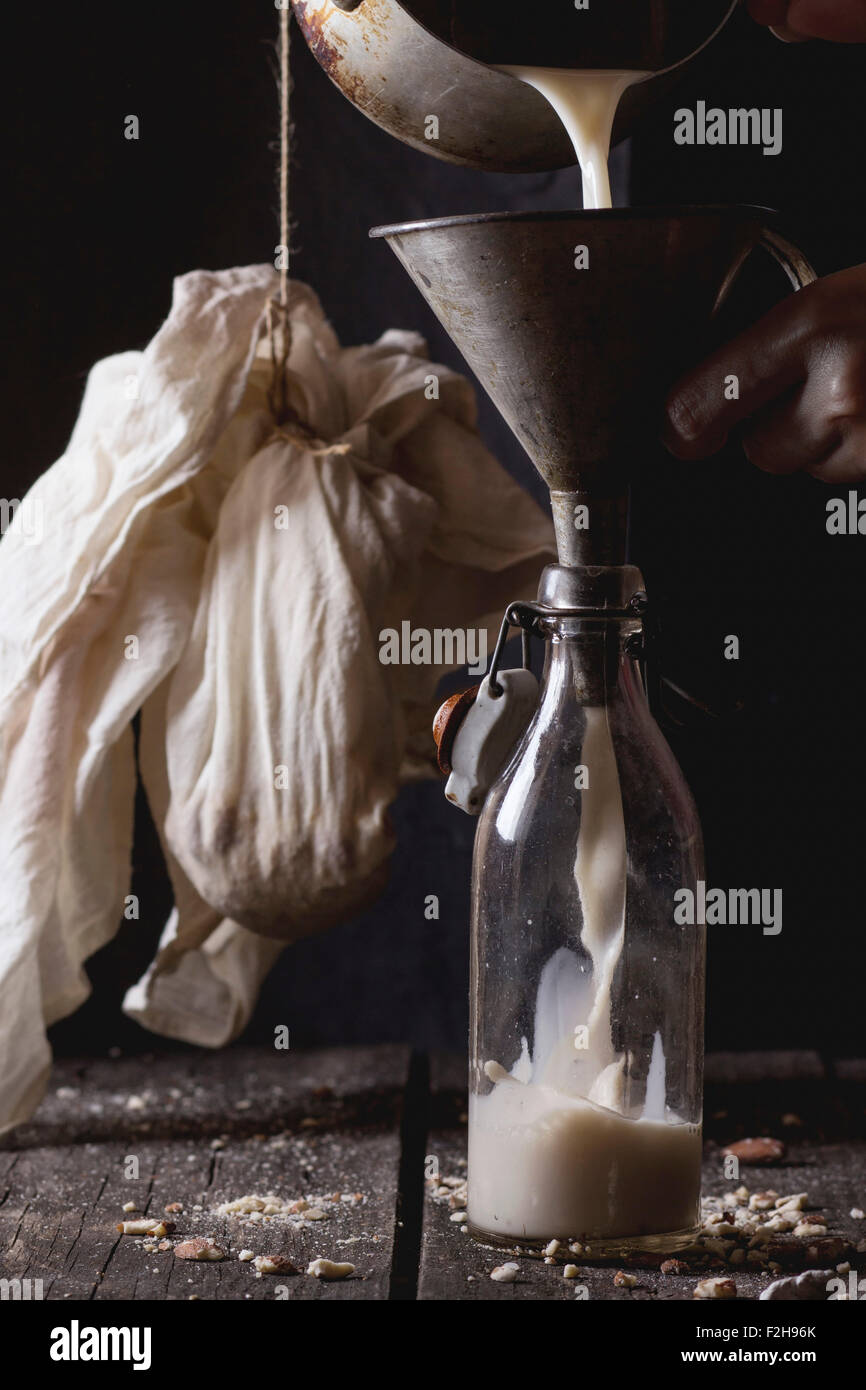 Process of making non-dairy almond milk - woman's hands pouring milk from pan in glass bottle via old funnel. Old wooden kitchen Stock Photo