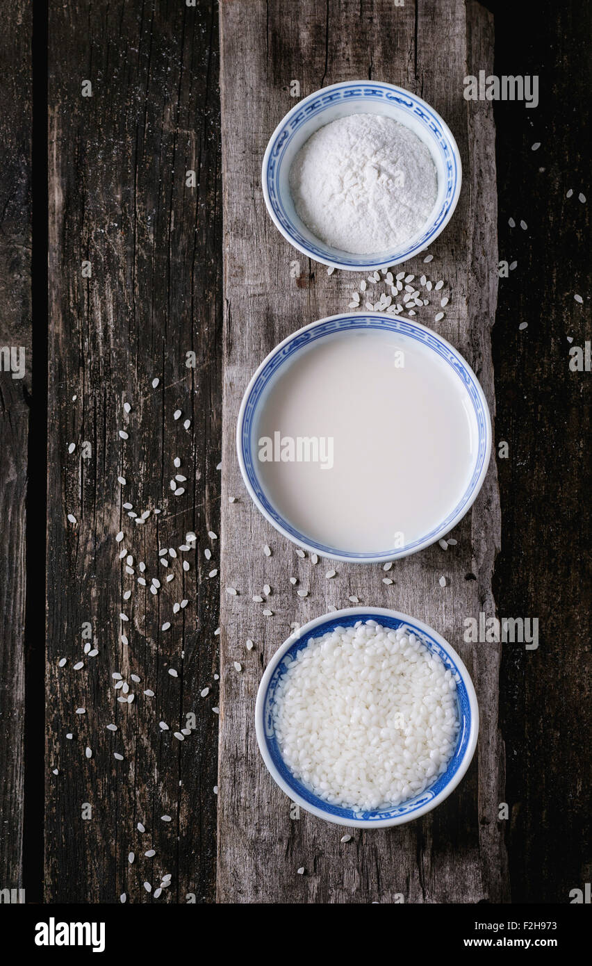 China style bowls with rice milk, rice flour and soaked rice over old wooden background with rice grains above. Top view. Stock Photo