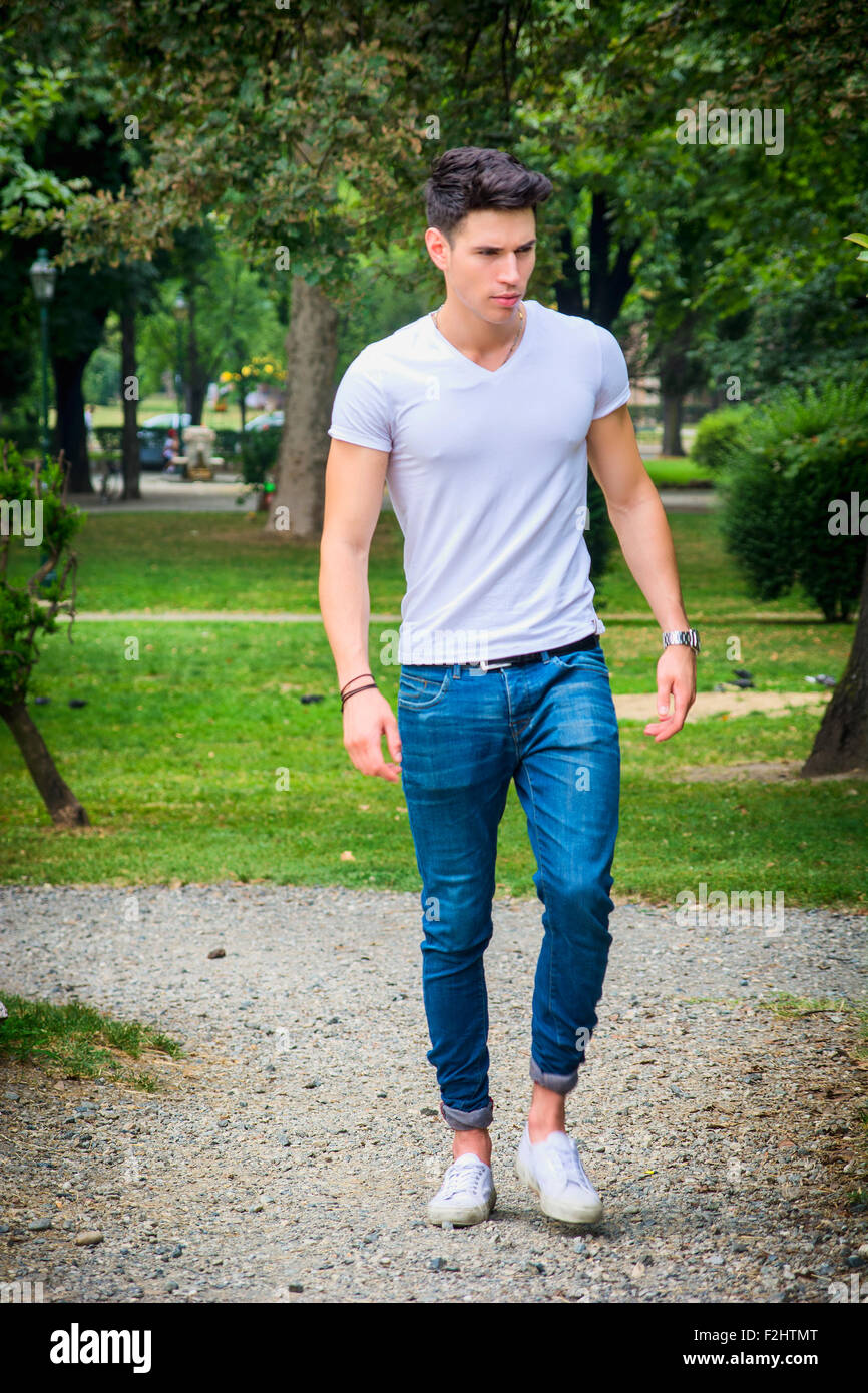 Handsome young man in white t-shirt and jeans outdoor in city park, walking toward camera, serious Stock Photo