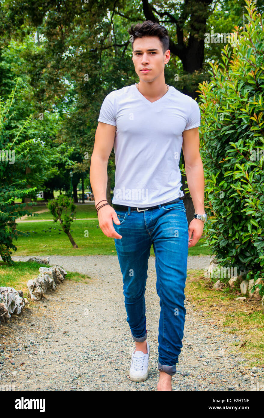 Handsome young man in white t-shirt and jeans outdoor in city park, walking toward camera, serious Stock Photo