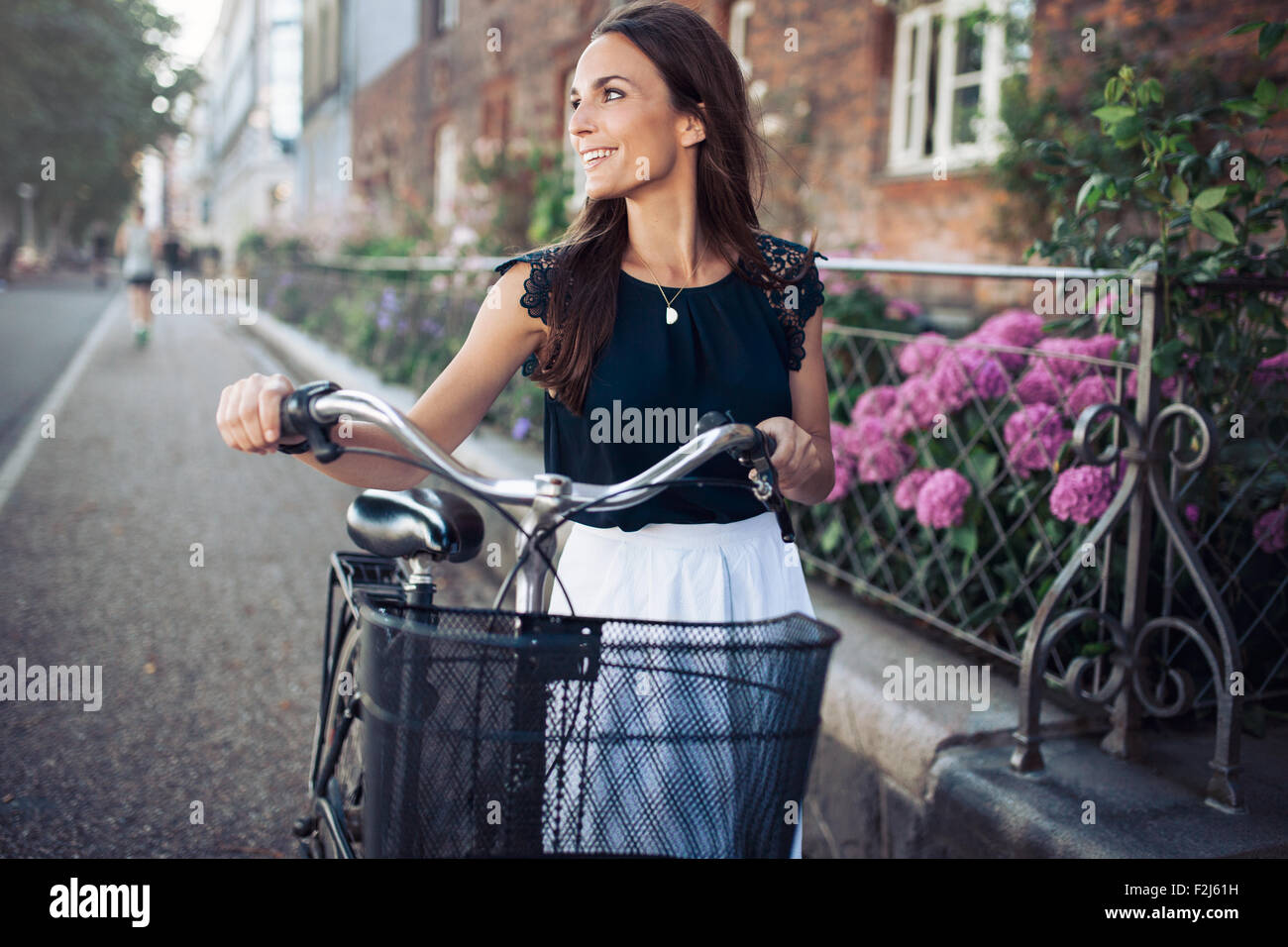 Cheerful young woman looking away while walking down the street with a bike. Female with a bicycle on city road looking at a vie Stock Photo