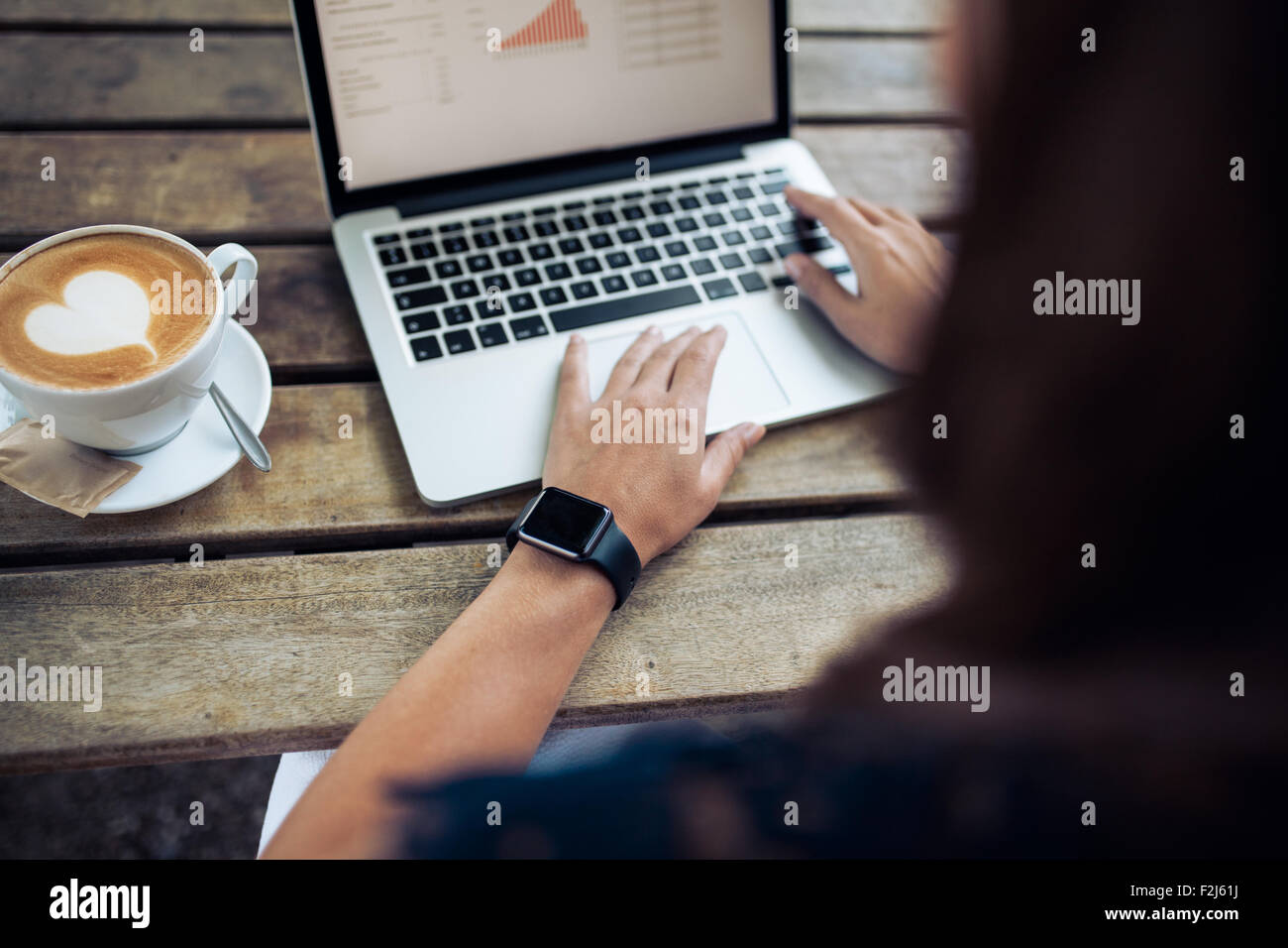 Woman typing on keyboard of a laptop with a coffee cup on wooden table. Female wearing a smartwatch at cafe. Stock Photo