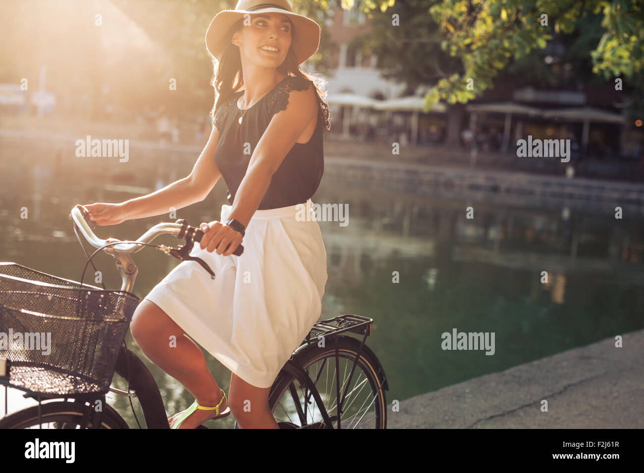 Portrait of beautiful young woman riding a bicycle along a street near pond on a summer day. Female wearing hat looking away whi Stock Photo