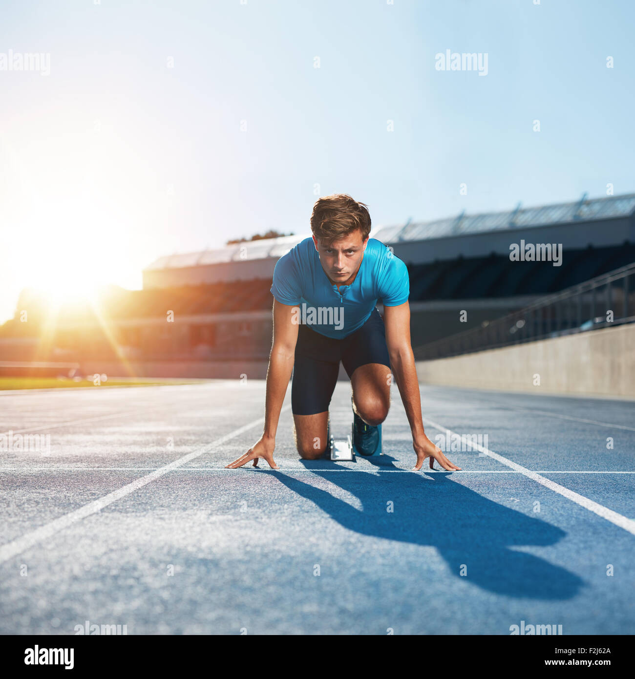 Fit and confident man in starting position ready for running. Male athlete about to start a sprint looking at camera with bright Stock Photo