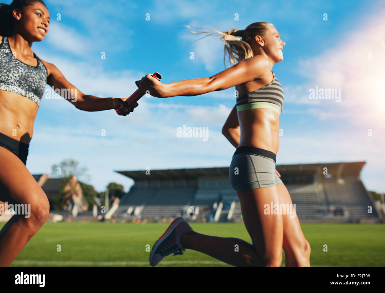 Female athletes passing over the baton while running on the track. Young women run relay race, track and field event. Stock Photo