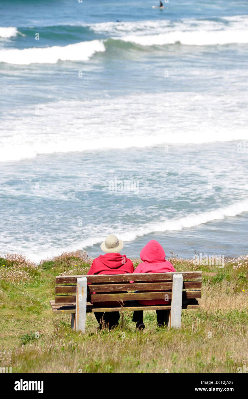 Two 'oldies' in red coats sitting on a cliff top seat - contemplating the incoming tide - white breakers - bright sunlight Stock Photo