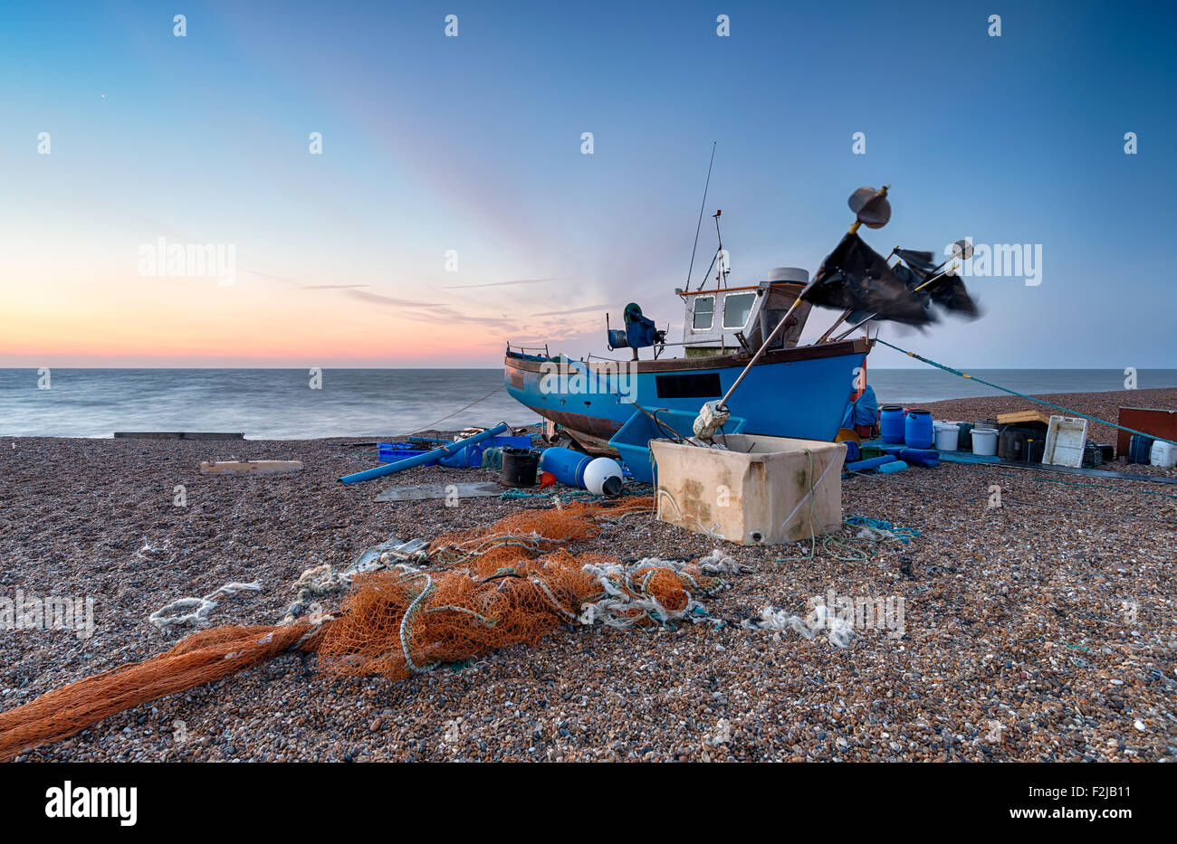 A blue fishing boat at sunrise with flags blowing in the breeze on a pebble beach at Aldeburgh in Suffolk Stock Photo