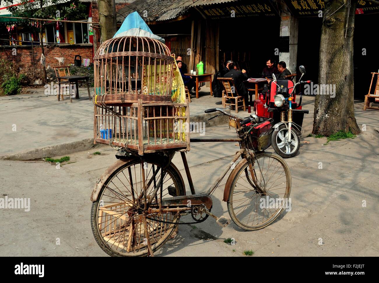 Long Feng, China:   Bicycle with two wooden bird cages strapped on the back parked near a group of men seated at tables Stock Photo