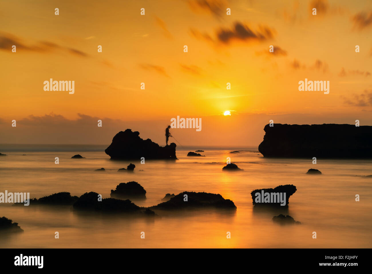 Sunset silhouettes create a beautiful long exposure photo of a man fishing from rocks on the stunning Indonesian island of Bali during sundown Stock Photo