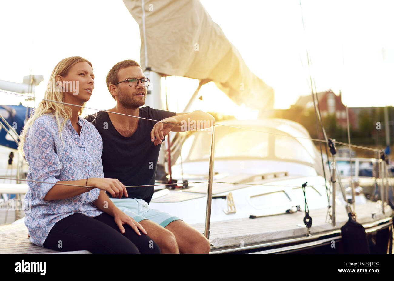 Sweet Young Lovers Sitting In Front of a Yacht and Looking Into the Distance During Sunset. Stock Photo