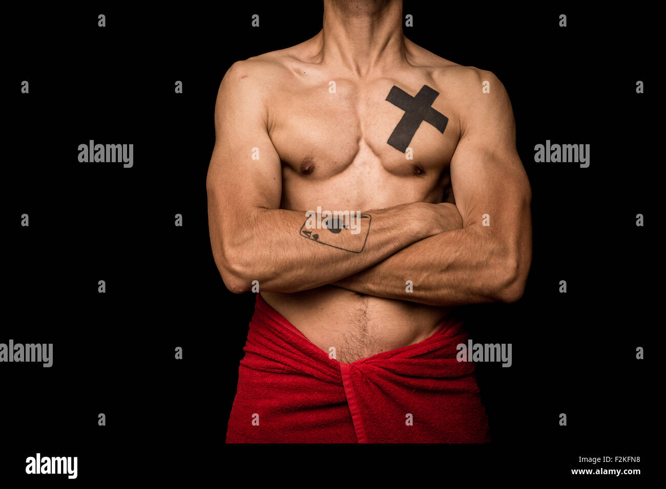 a fit, tattooed young man stands against a black background with a red towel around his waist, displaying his muscles Stock Photo