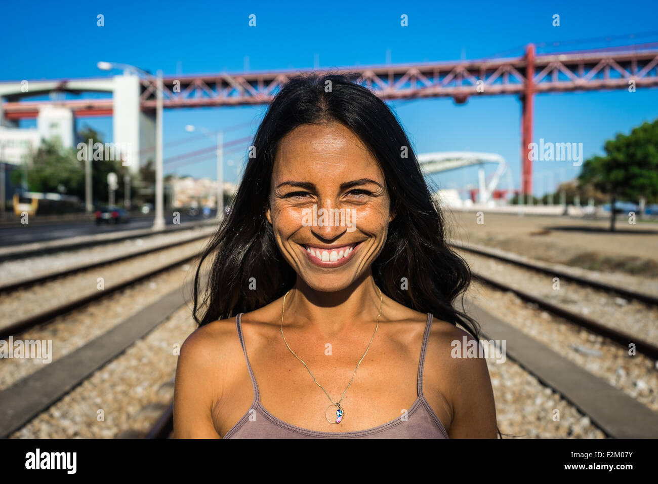 Pretty Portuguese girl smiles on vacant train tracks in front of the Abril de 25 Bridge in Lisbon, Portugal. Stock Photo