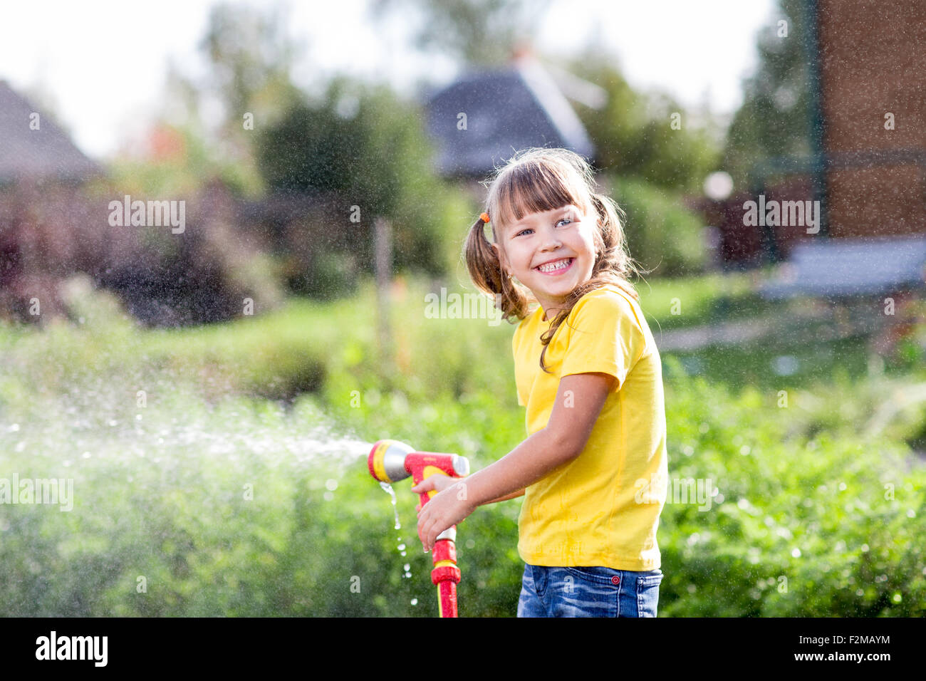 Cheerful child watering plants from hose spray in garden at backyard of house at sunny summertime Stock Photo