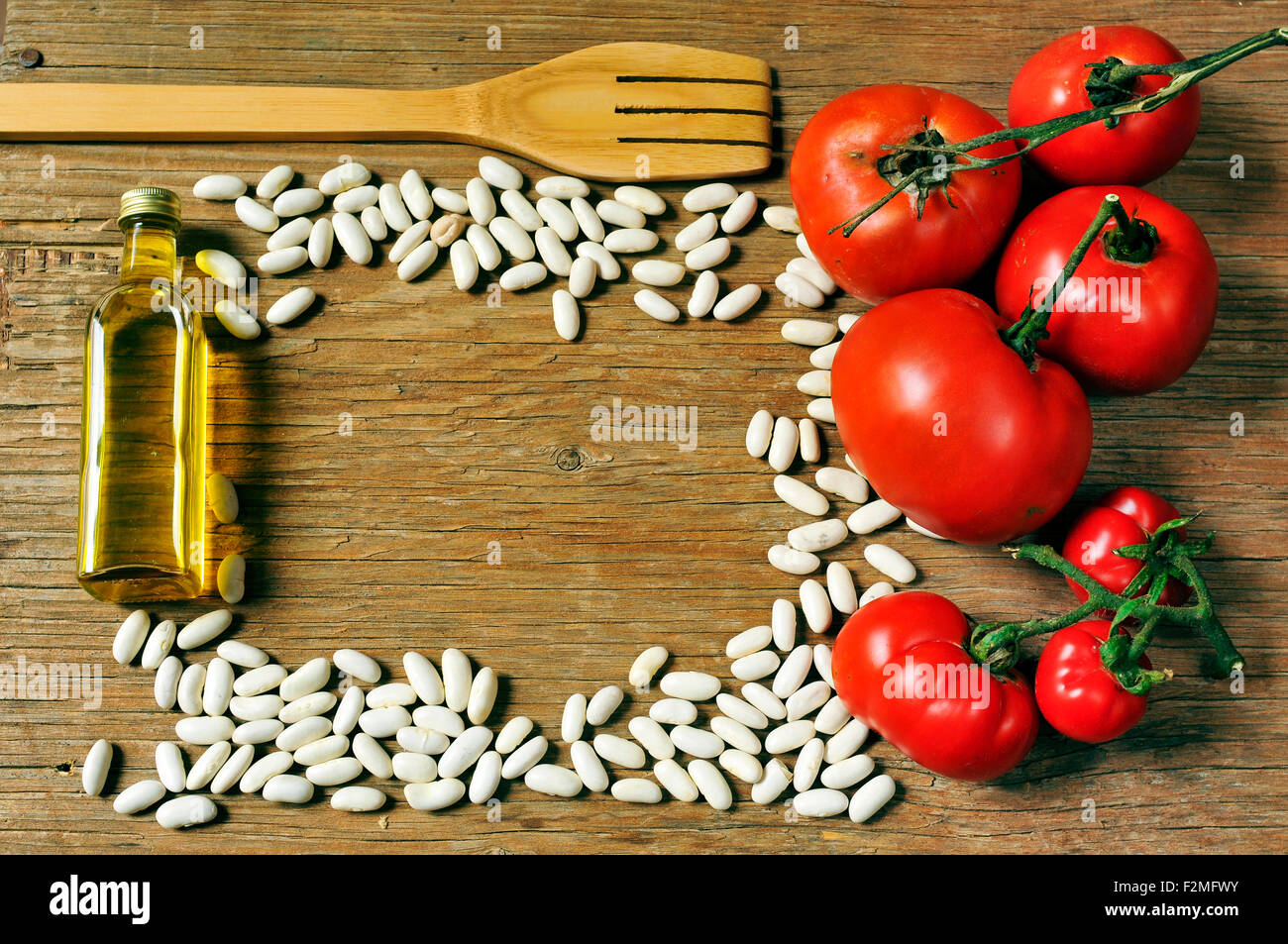 high-angle shot of a wooden fork, a bottle of olive oil, some white beans and some tomatoes on a rustic wooden table arranged to Stock Photo