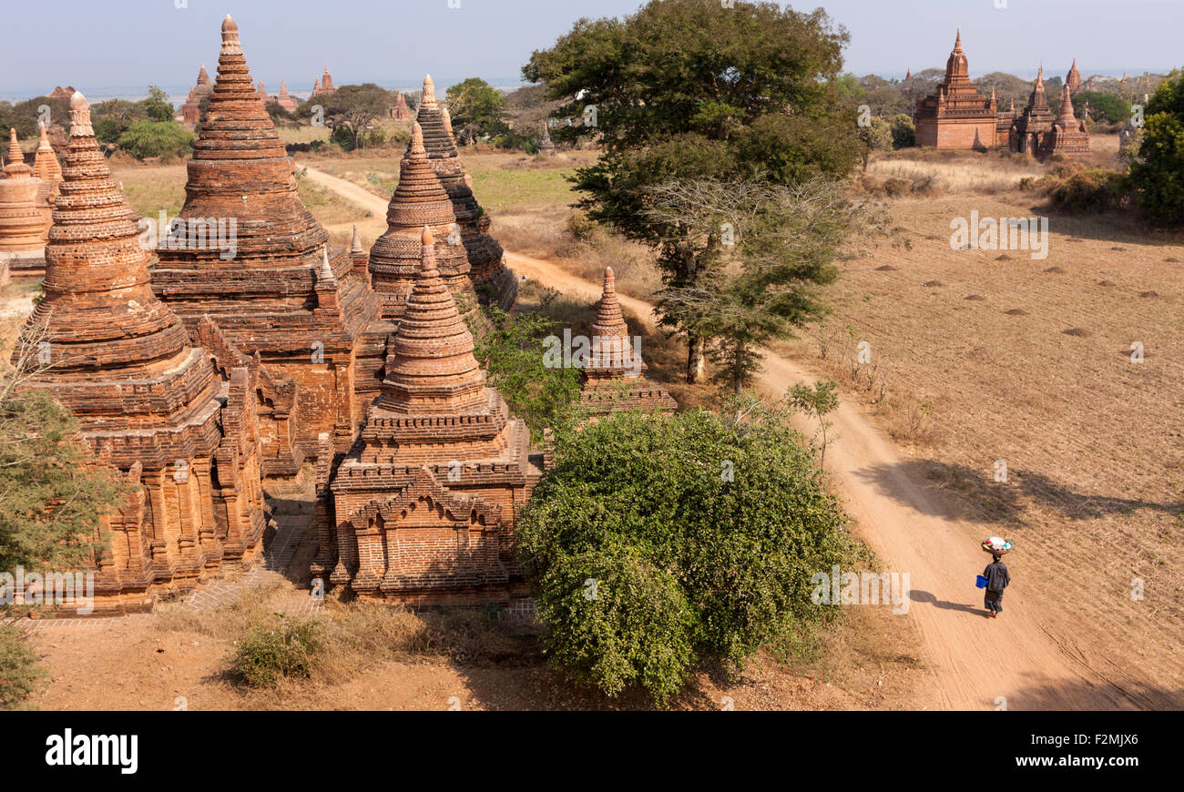 View from Kay Min Gha pagoda at Bagan Myanmar. A woman walking on dirt path between pagodas carries a large basket on her head. Stock Photo