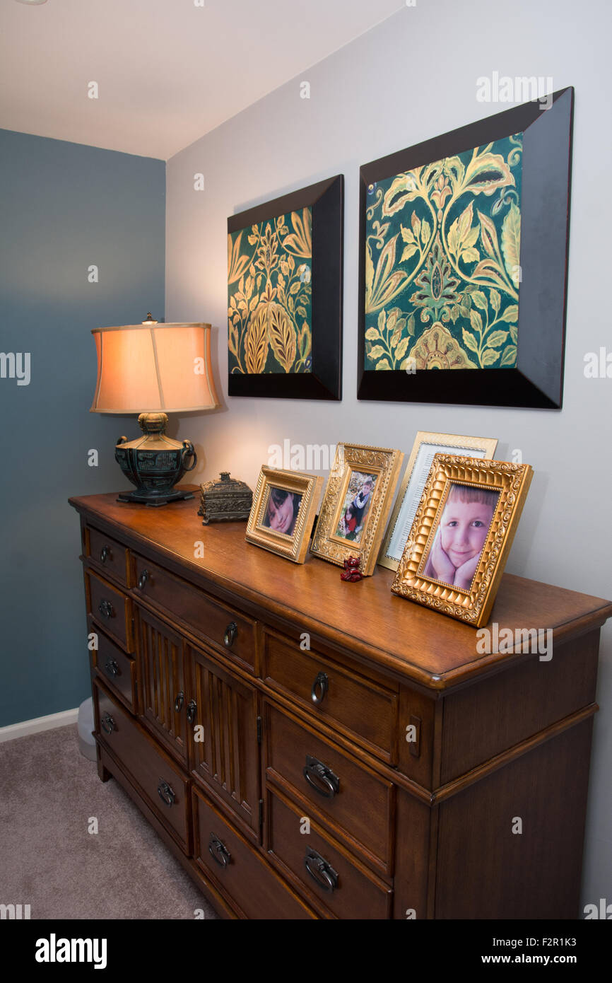 A dresser in a bedroom with art above and a lamp and framed photographs sitting on it. Stock Photo