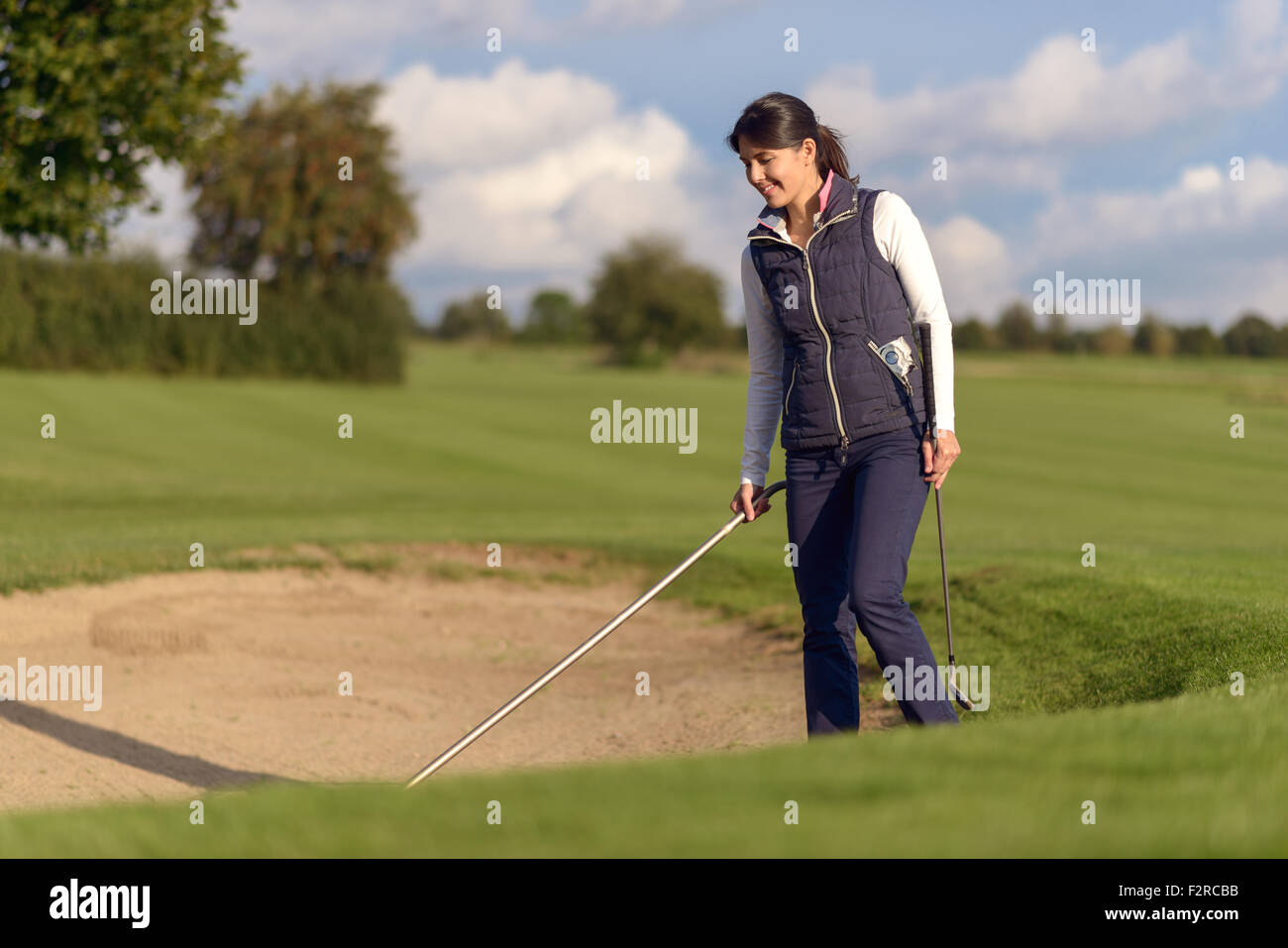 Woman golfer raking a sand bunker on a golf course to level off the surface after she has played her shot to retrieve her ball Stock Photo