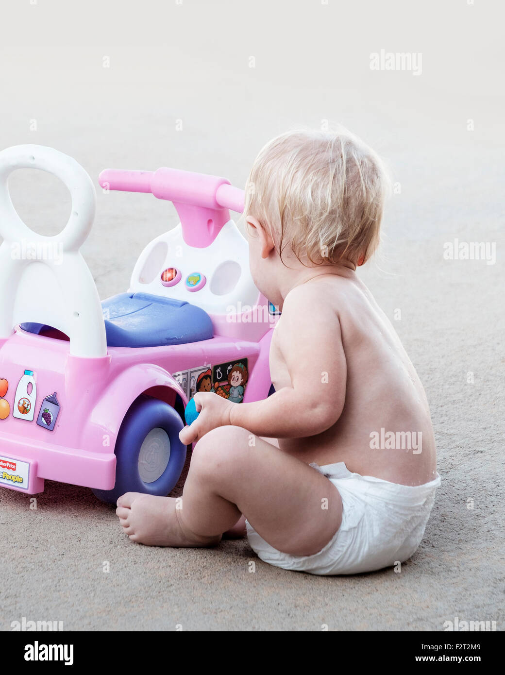A one year old blonde Caucasian baby girl in a diaper only plays with her riding toy car. Stock Photo
