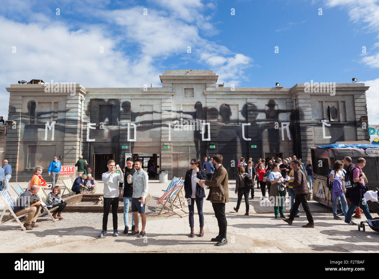 Visitors inside Banksy's Dismaland stand in front of a building with the word Mediocre painted on it by Axel Void. Stock Photo