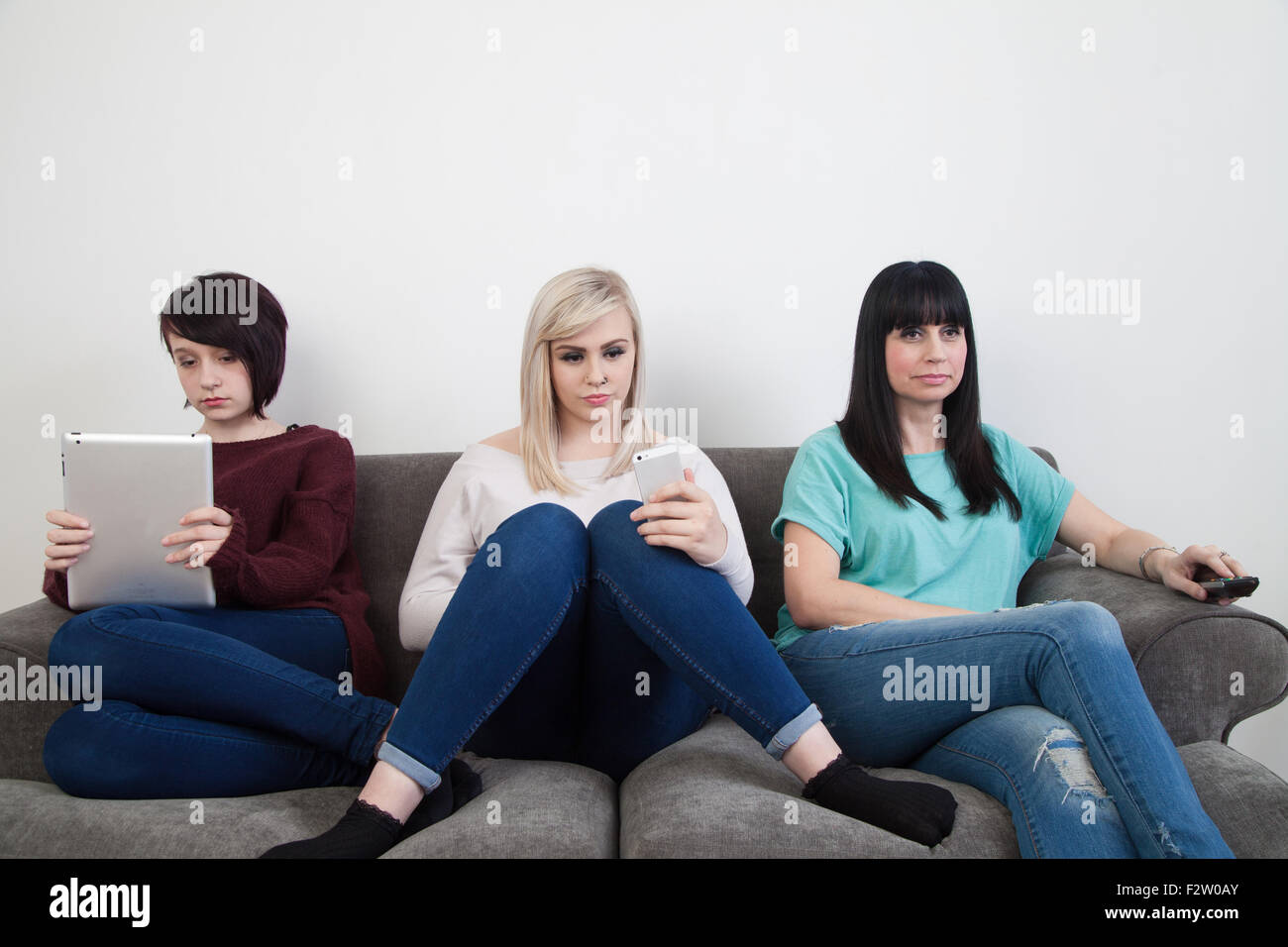 Mother and her two daughters sitting on a sofa watching TV , using an ipad and iphone. Stock Photo