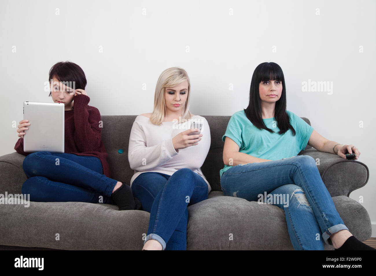 Mother and her two daughters sitting on a sofa watching TV , using an ipad and iphone. Stock Photo