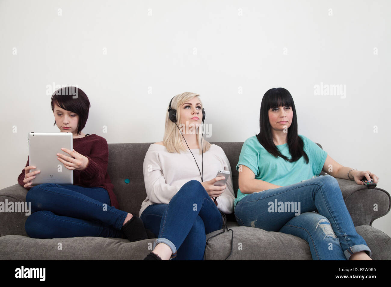 Mother and her two daughters sitting on a sofa watching TV , using an ipad and listening to music. Stock Photo