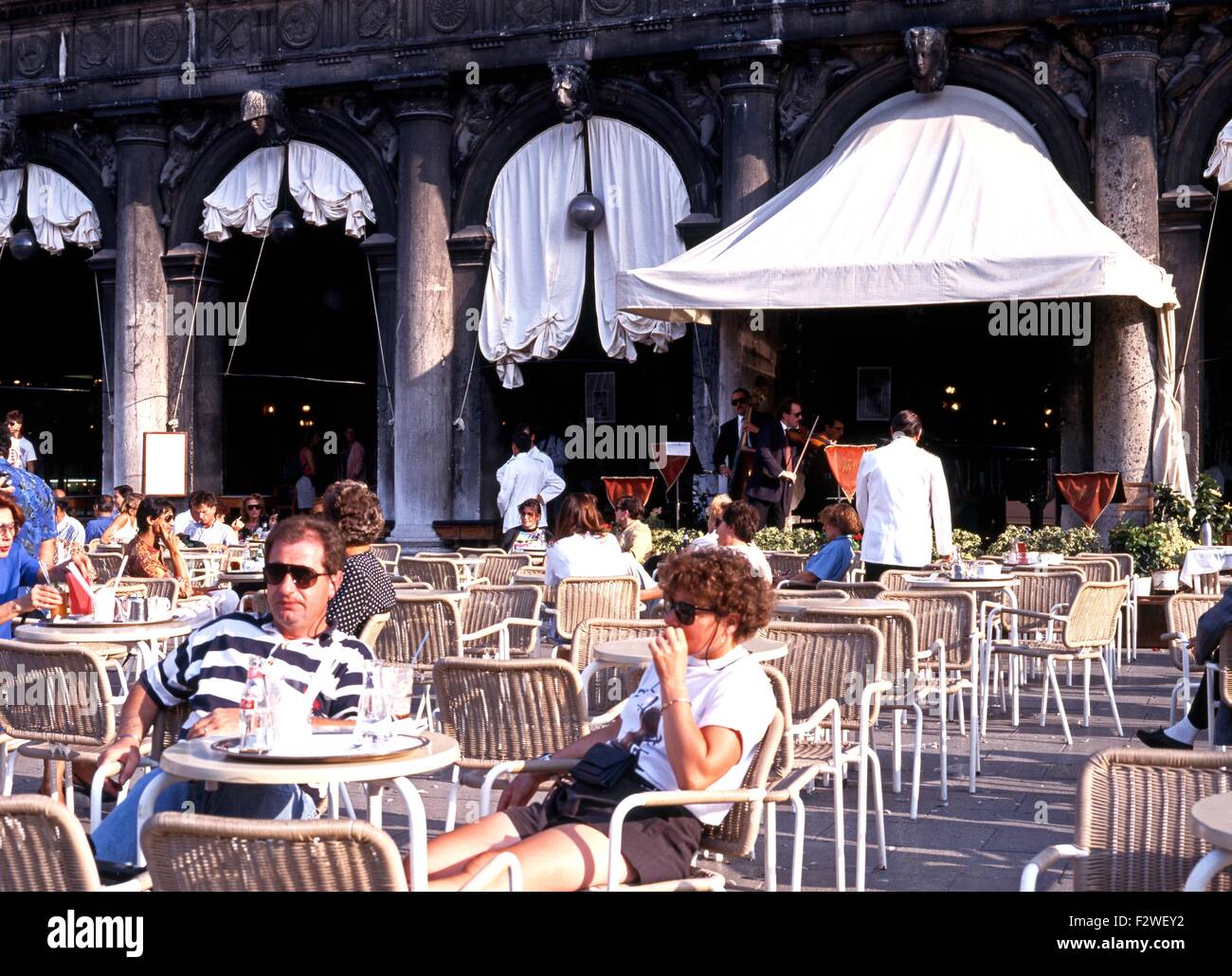 Tourists at a pavement cafe with musicians to the rear in St Marks Square, Venice, Veneto, Italy, Europe. Stock Photo