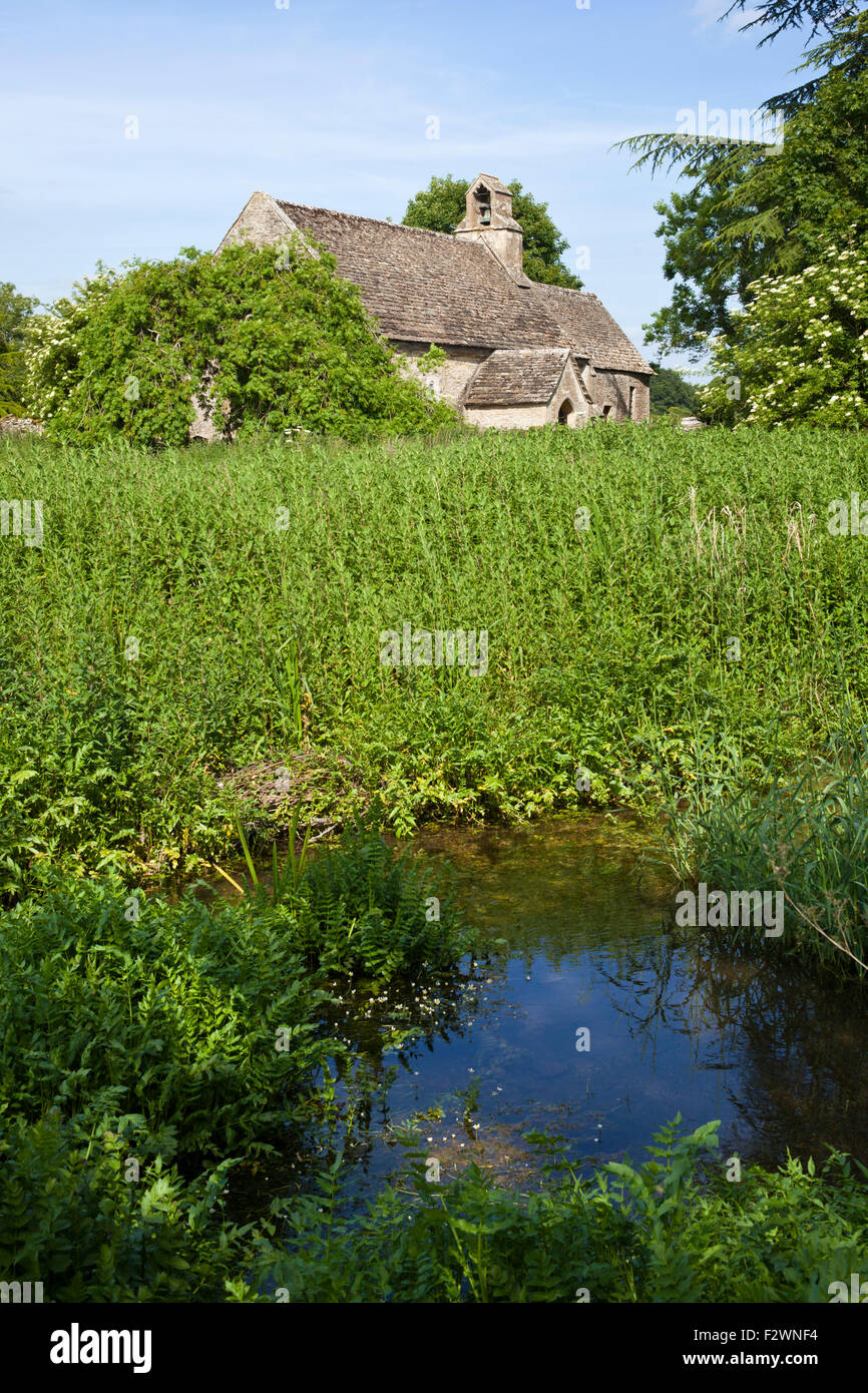 The Ampney Brook flowing past the Norman church of St Mary in the Cotswold village of Ampney St Mary, Gloucestershire UK Stock Photo