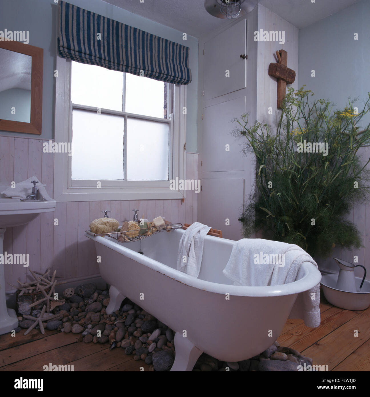 Striped blind on frosted window in nineties bathroom with large group of seashore pebbles below roll top bath Stock Photo