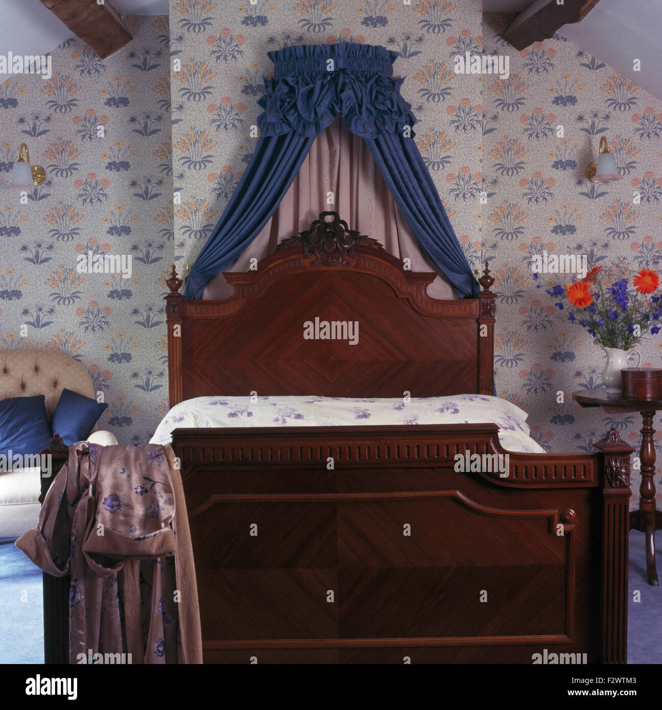 with blue drapes above carved wooden bed in bedroom with floral wallpaper Stock