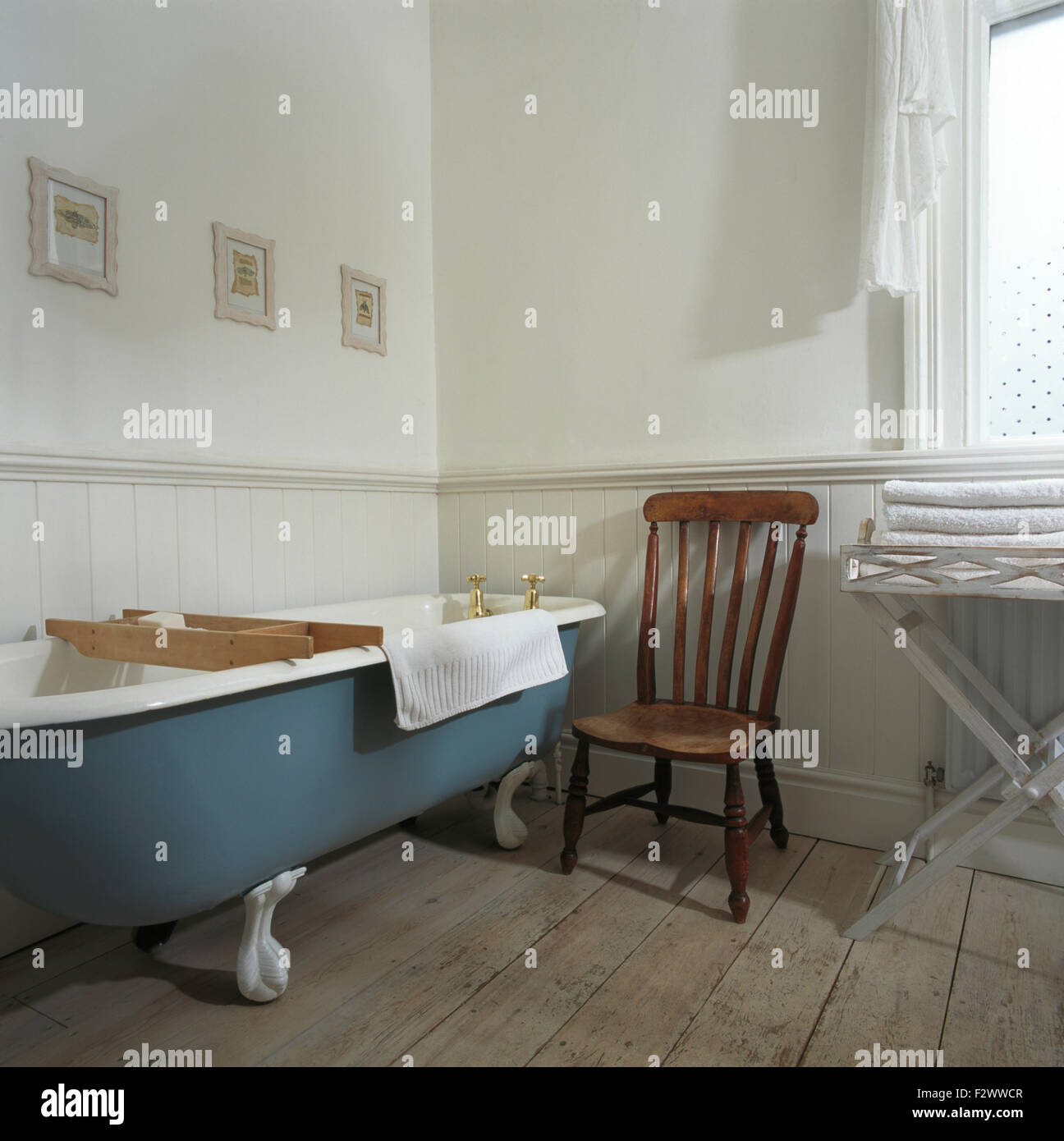 Antique wooden chair beside blue roll top bath in traditional bathroom with lime washed floor boards Stock Photo