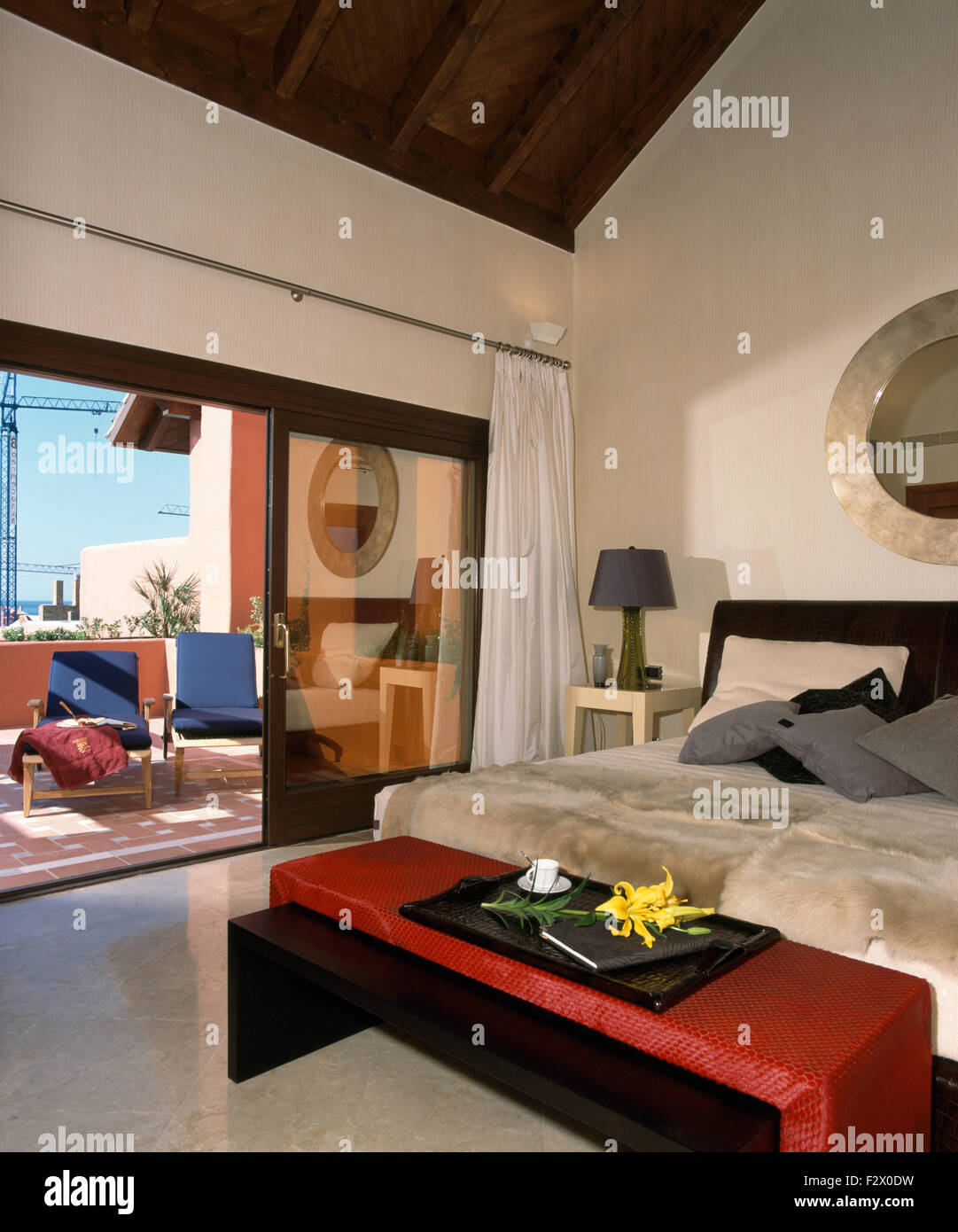 Red stool below bed with faux fur throw in modern Spanish bedroom with open glass doors to patio Stock Photo