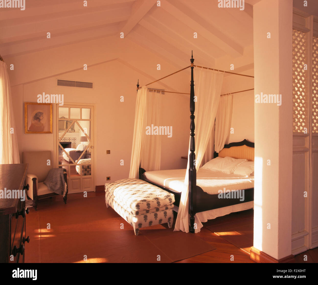 Upholstered ottoman stool below four poster bed with white drapes in white Spanish coastal