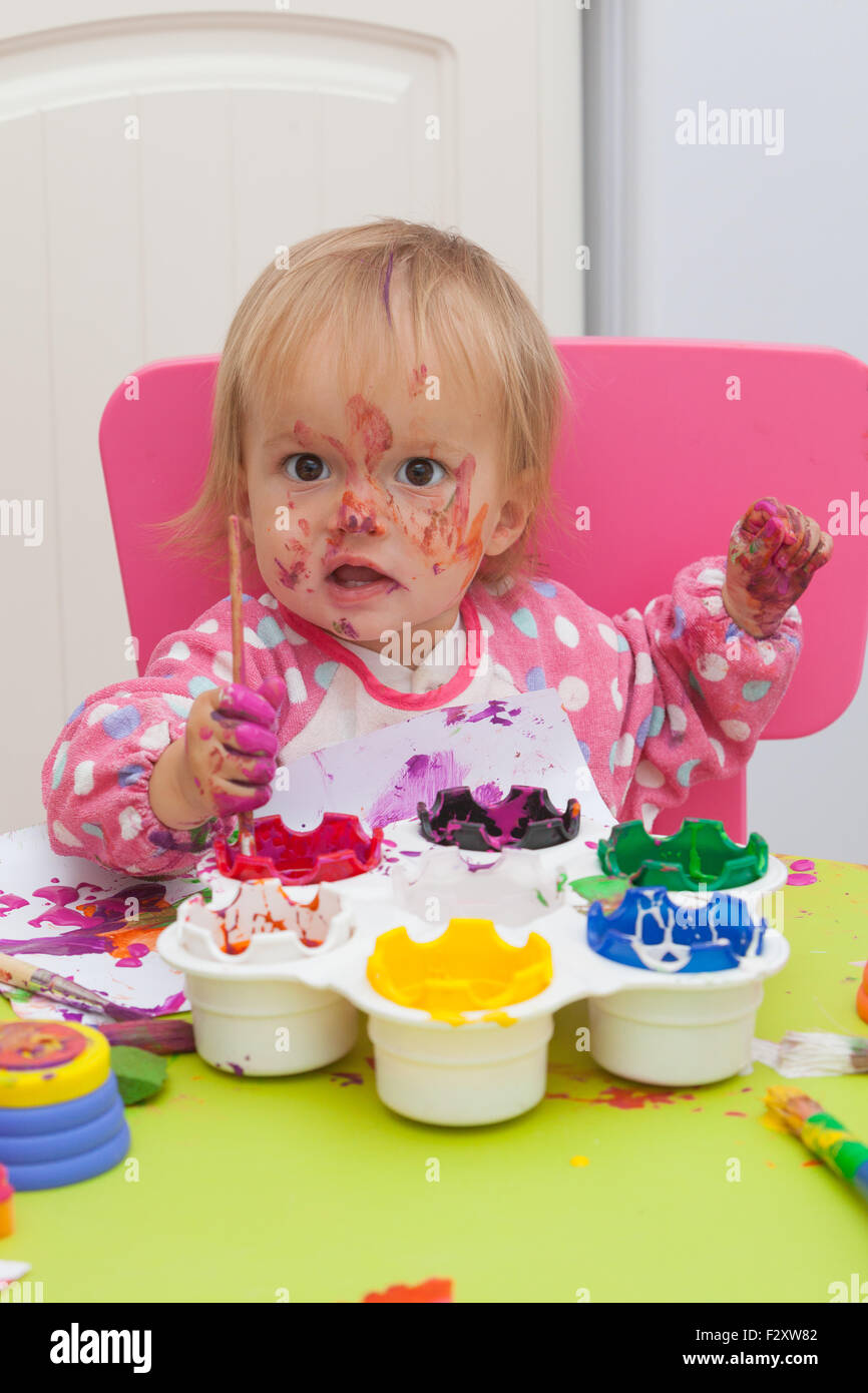 A 16 month old, blond haired, caucasian, girl playing with paints and a paint brush while sitting inside at a small table. Stock Photo