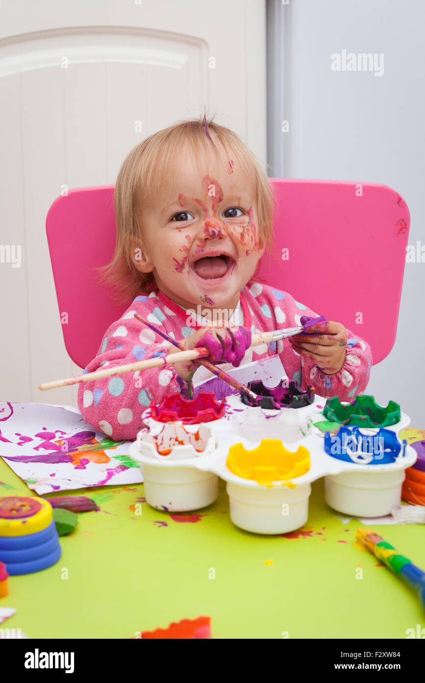 A 16 month old, blond haired, caucasian, girl playing with paints and a paint brush while sitting inside at a small table. Stock Photo