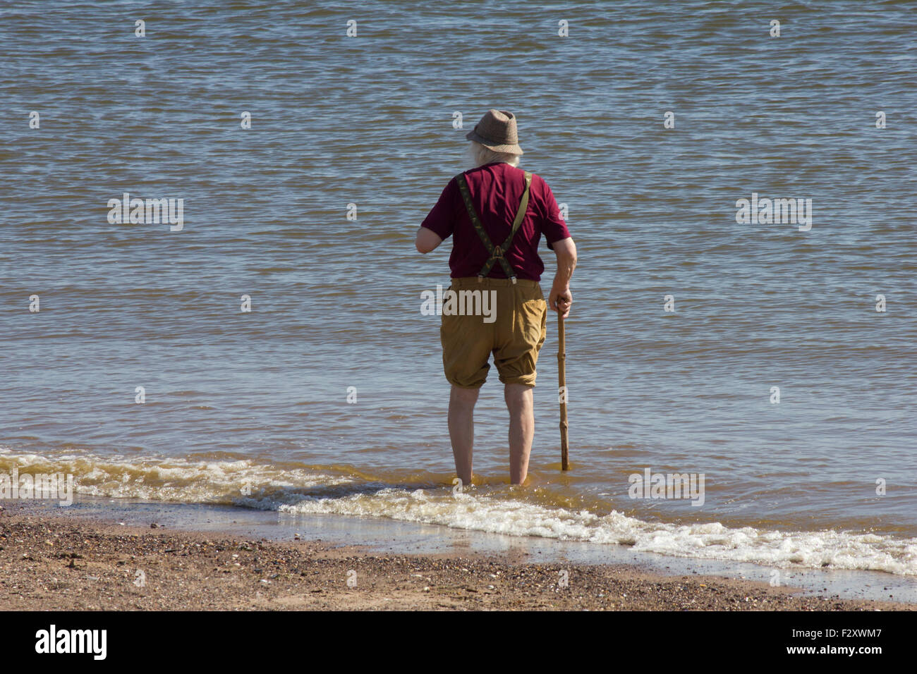 An elderly man wearing a hat carrying a walking stick with his trouser legs rolled up paddling in the sea Stock Photo