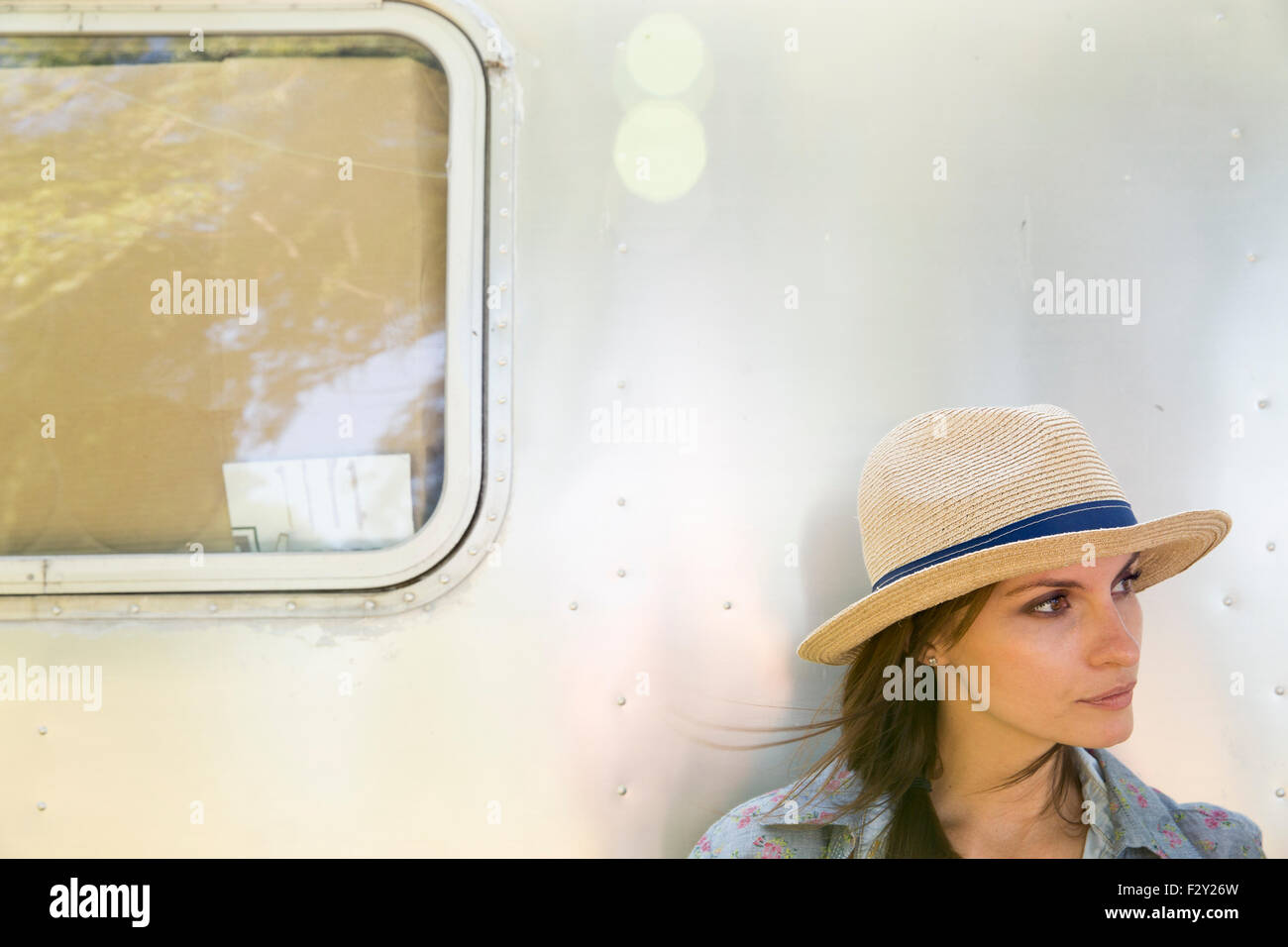 A young woman wearing a hat sitting in the shade of a silver coloured trailer. Stock Photo