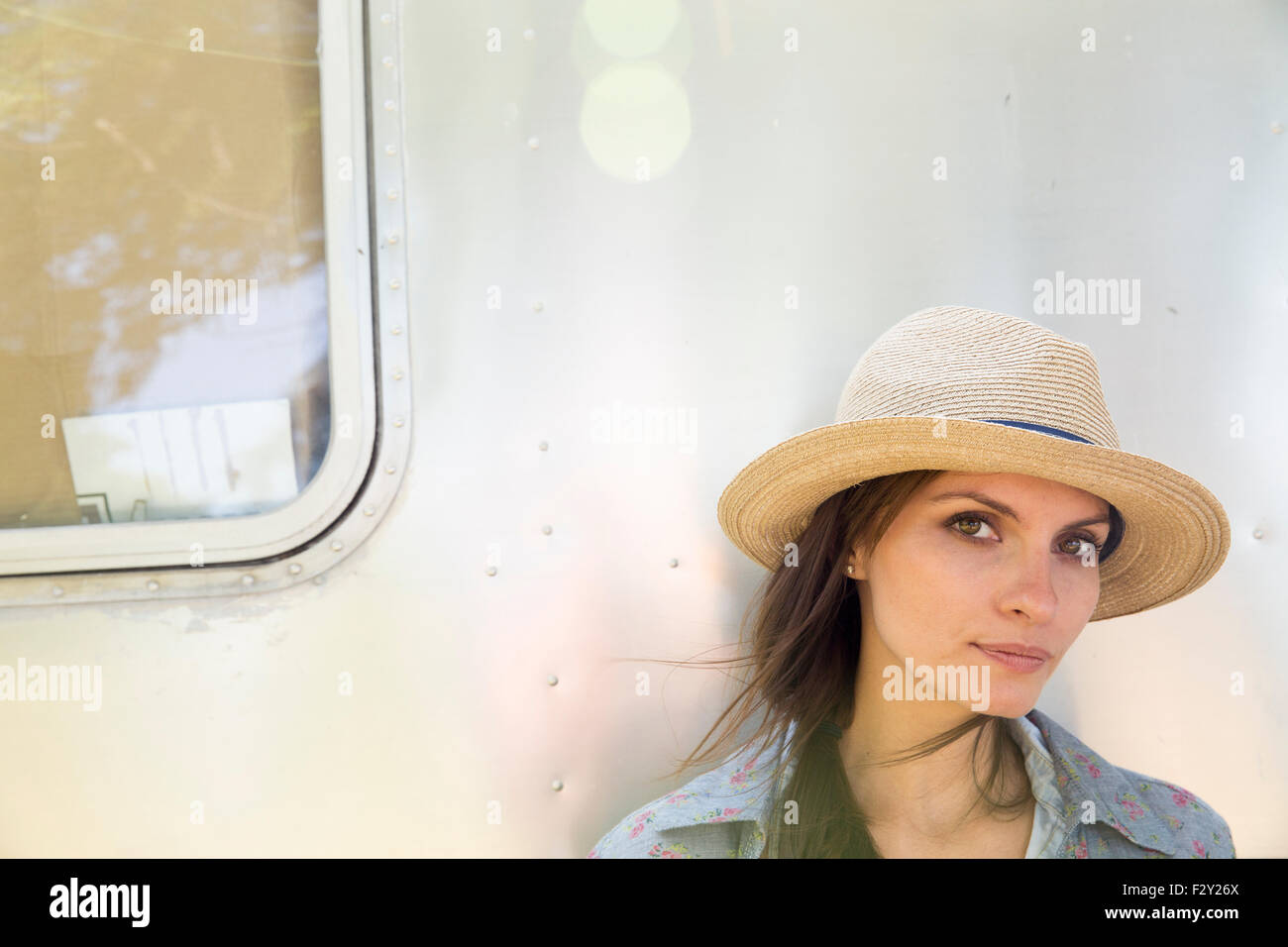 A young woman wearing a hat sitting in the shade of a silver coloured trailer. Stock Photo