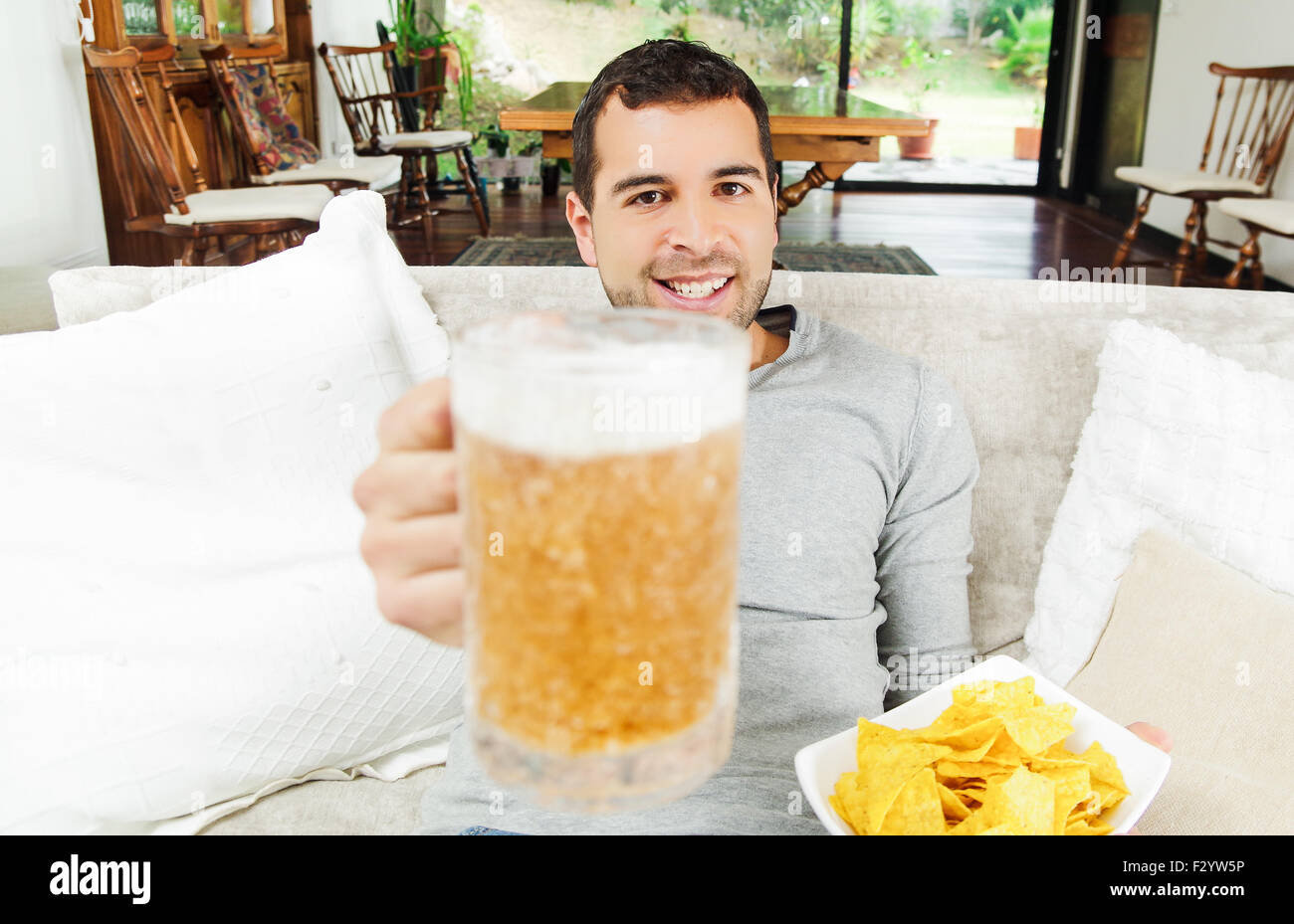 Hispanic pleased male wearing light blye sweater enjoying potato chips and beer sitting in white livingroom sofa facing camera Stock Photo