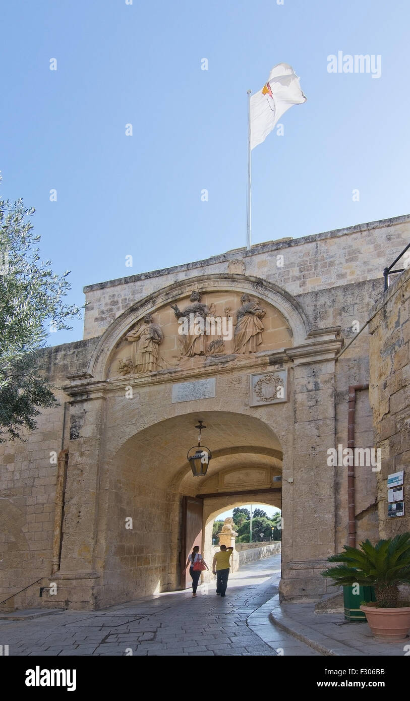 Arched entrance portal with flag, people coming through and old city walls on a sunny day in Mdina, Malta. Stock Photo