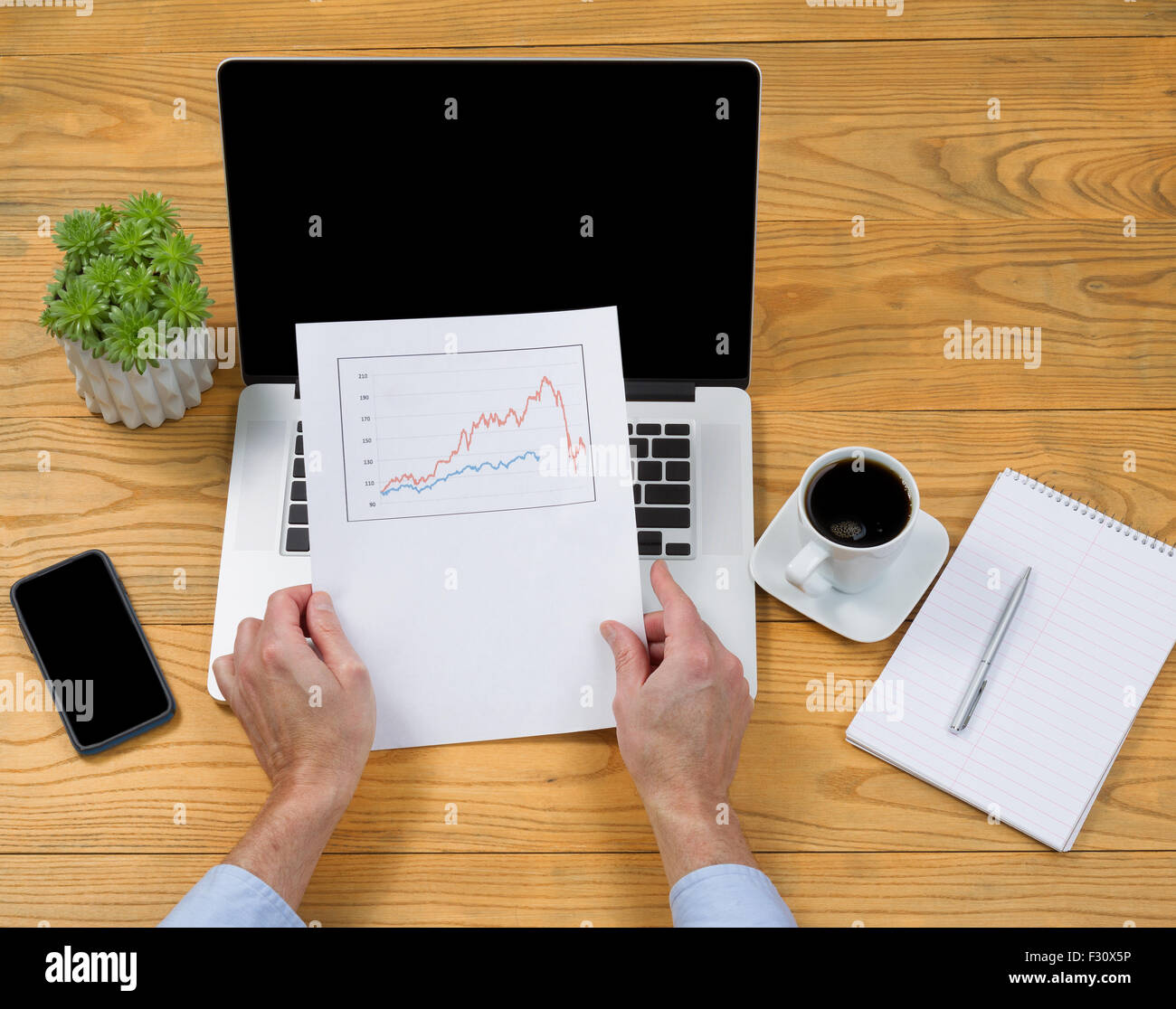 High angled view of male hands holding data graph with computer, coffee, notepad, pen and plant on desktop. Stock Photo