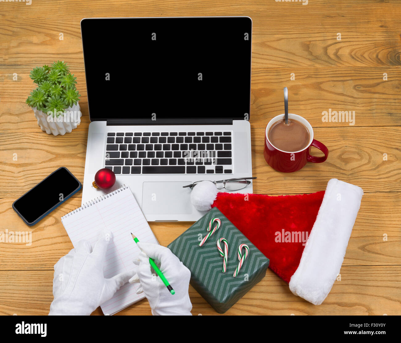 High angled view of Santa Claus writing his gift list with hot chocolate, computer, present, cap, notepad, pencil, cell phone, r Stock Photo